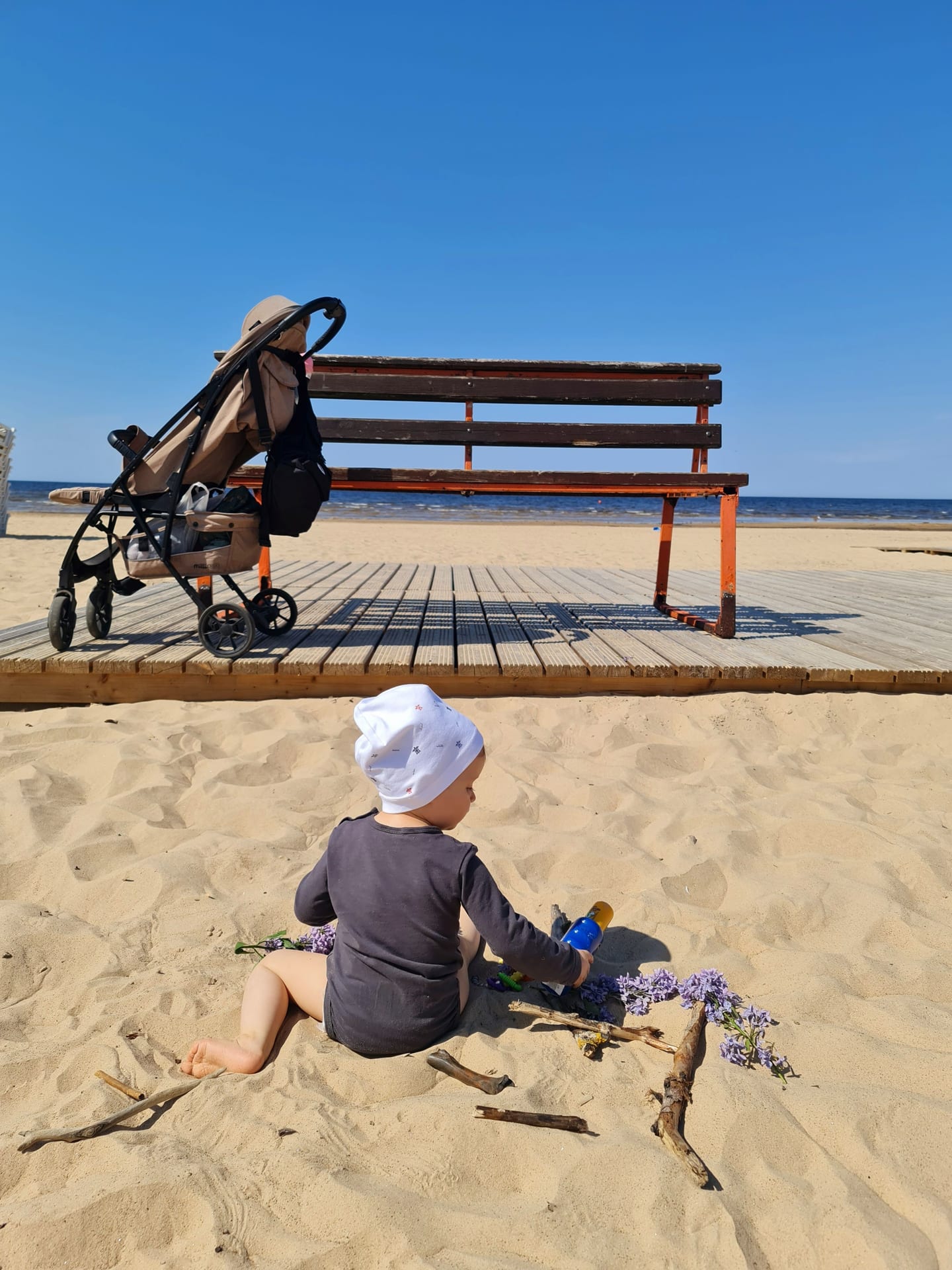 A baby plays on a sandy beach with a stroller and bench, perfect for summer vacation themes.